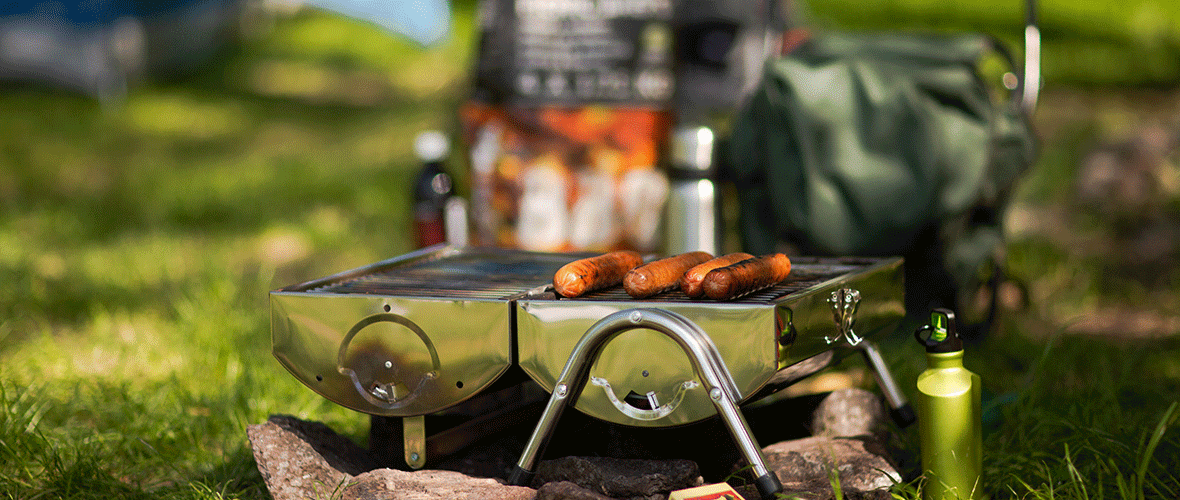 Portable camping grill with sausages being cooked outdoors at a campsite.