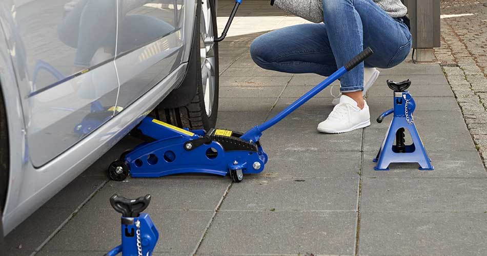 Person using a jack to lift a car surrounded by trestles on a tiled surface.
