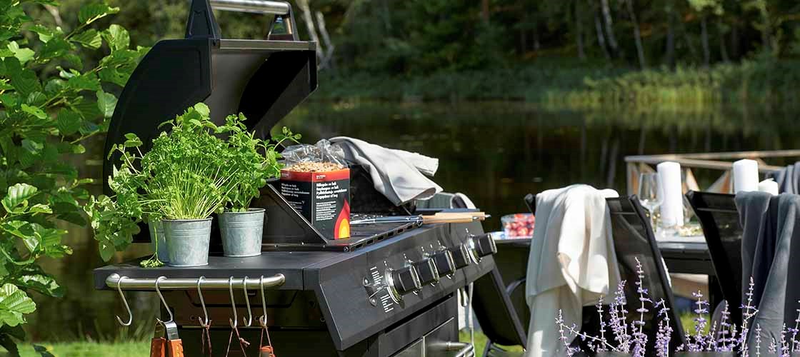 Outdoor gas grill by a lake, surrounded by greenery. Potted herbs sit on the side shelf of the grill. In the background, a set table with chairs is visible in a garden setting.