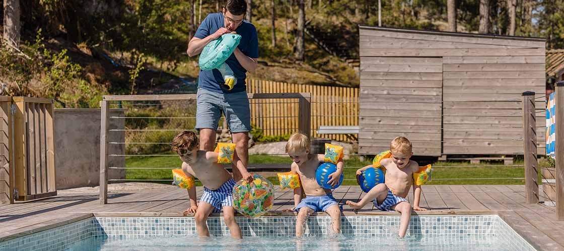 Flere personer sitter og står ved kanten av et utendørs svømmebasseng. Tre barn dypper føttene i bassenget, og en voksen mann står i bakgrunnen og blåser opp en badering. I bakgrunnen er en treterrasse, et gjerde, et gressområde og en liten bygning omgitt av trær. Bildet viser et sommermiljø der folk er ved et basseng for å svømme og slappe av.