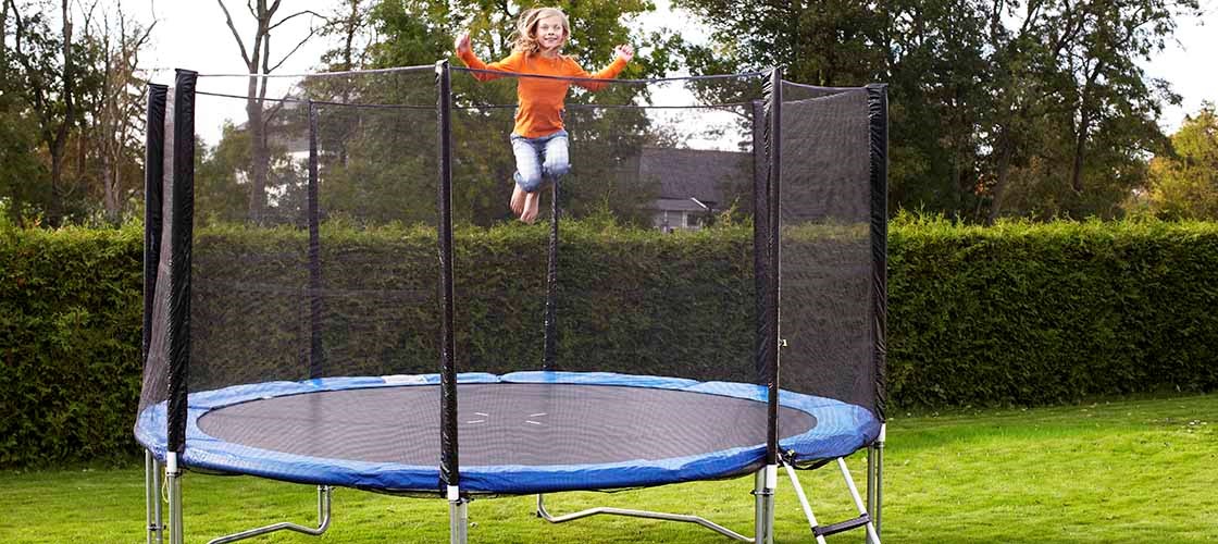 En person hopper på en rund trampoline med et sikkerhetsnett som er plassert på en plen i en hage. Trampolinen har en stige på siden og er omgitt av hekker og trær i bakgrunnen. Bildet viser et utendørsmiljø med fokus på bruk av en trampoline med sikkerhetsnett for økt sikkerhet.