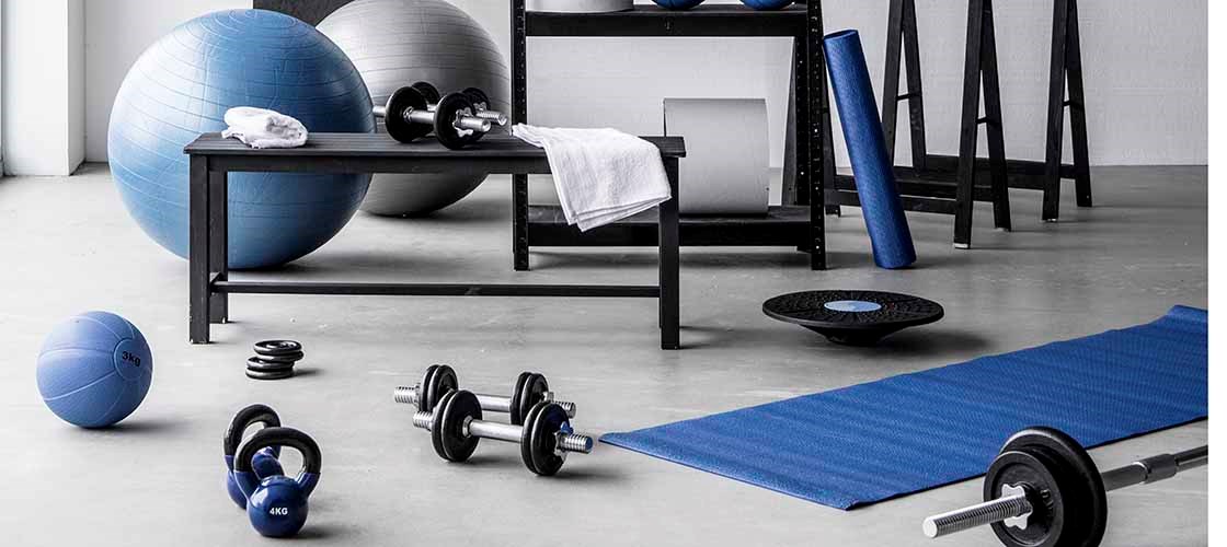 Et treningsrom med blått og hvitt treningsutstyr, inkludert yogamatte, manualer, kettlebell, medisinballer og store treningsballer. En svart hylle inneholder mer utstyr som vekter, håndklær og treningsstrikker. På veggen vises en håndtegnet oversikt med teksten «Squat Challenge».