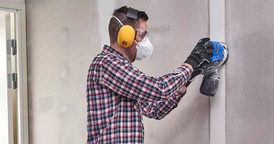 En mann iført rutete arbeidsskjorte, arbeidshansker, hørselsvern og ansiktsmaske sliper gipsvegg med en batteridrevet, blå slipemaskin.