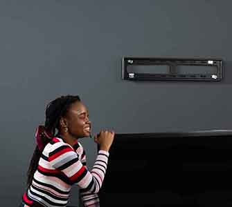 En kvinne i en hvit og rødstripet genser løfter den ene siden av en TV. Hun ler og har håret satt opp i en knute. På den grå veggen i bakgrunnen er et veggmontert TV-stativ synlig.
