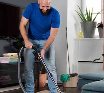 A man in a blue t-shirt and blue jeans is vacuuming in a living room. Green plants stand in the window and behind him a TV is visible on the wall.
