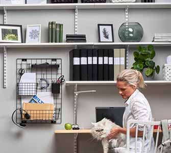 On the left of the picture, a woman in a white shirt sits holding a white cat in her lap. Behind her, the gray wall is filled with shelves filled with binders, plants, and other office furnishings.
