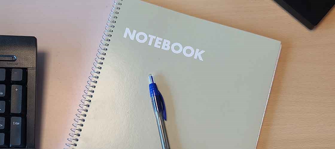Spiralnotatbok med teksten «NOTATBOK» og blå kulepenn på skrivebordet.
